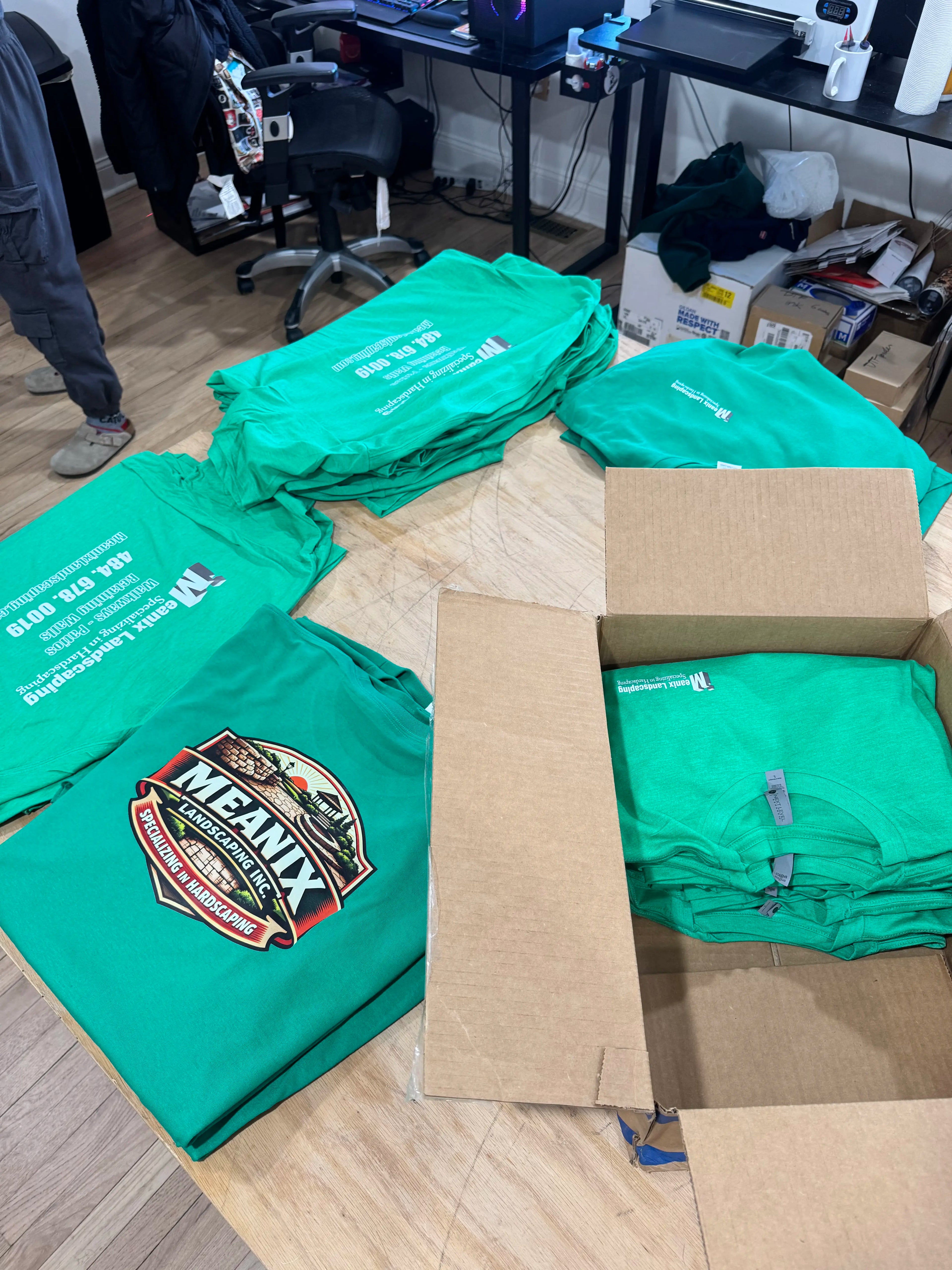Green t-shirts with a logo on a wooden floor, with a cardboard box and more t-shirts in the background.