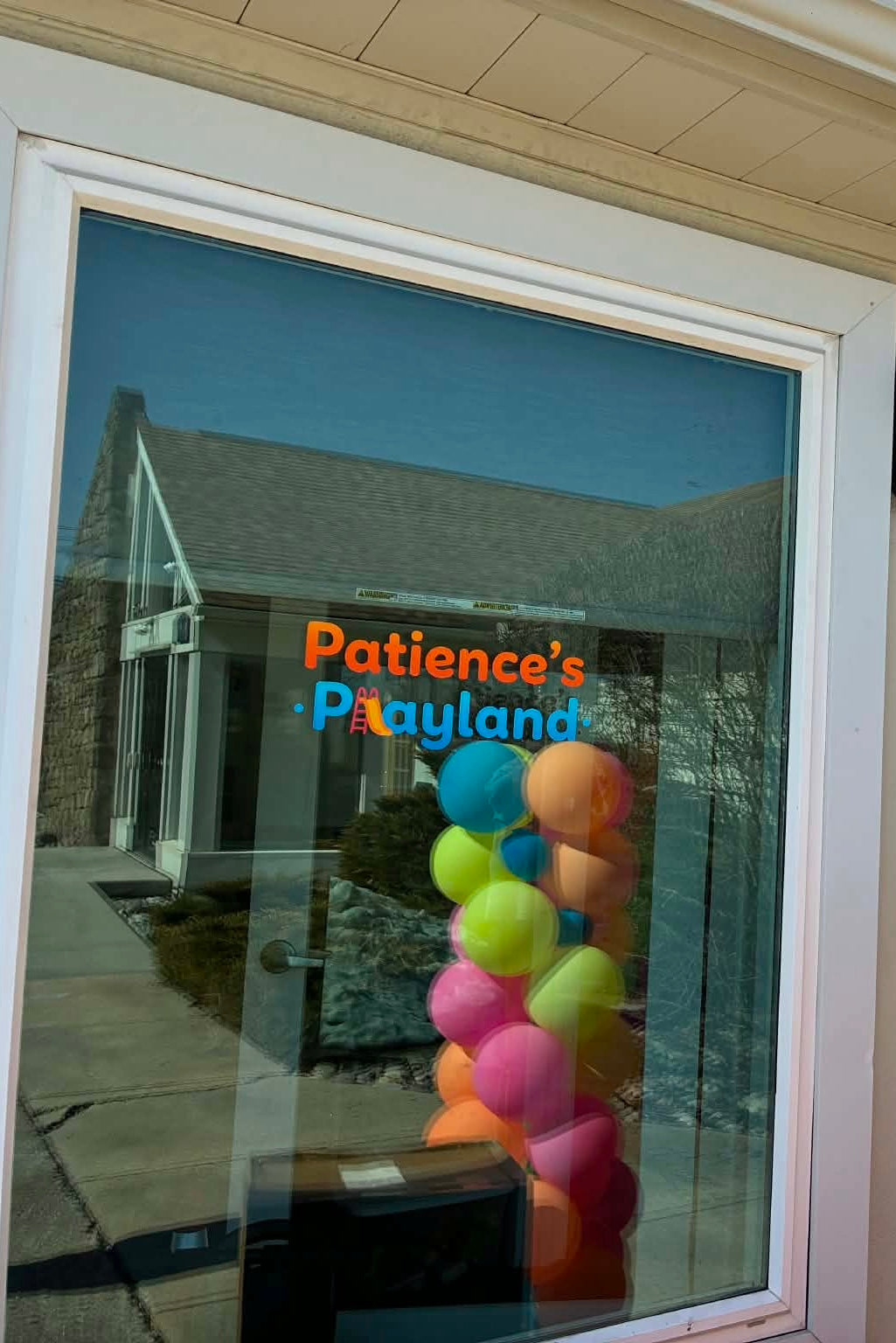 Glass door with colorful balloons and 'Patience's Playland' sign reflected on it.